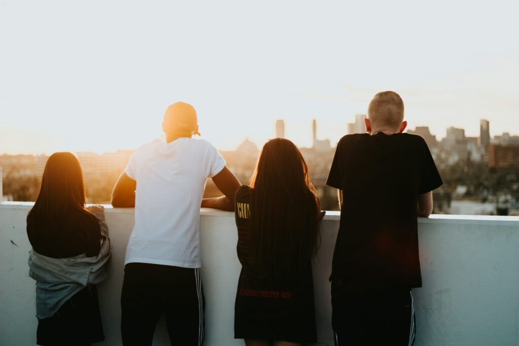 Auf dem Bild sind vier Personen zu sehen, die an einer Mauer gelehnt auf eine Stadt schauen hinter der die Sonne untergeht.